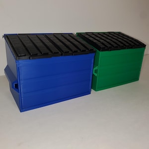 May include: Two miniature dumpsters, one blue and one green, are side-by-side. Each has a black, ridged lid and a rectangular shape. The blue dumpster has a small, rectangular opening on the side. The green dumpster has a handle on the side.