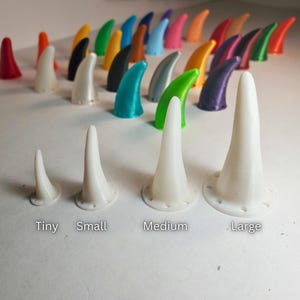 Op de afbeelding: Een maattabel toont diverse kleurrijke, hoornvormige objecten. De tekst "Size Guide" staat bovenaan. Vier witte hoornvormige objecten zijn gelabeld met "Tiny", "Small", "Medium" en "Large". De achtergrond toont rijen kleurrijke hoorns.
