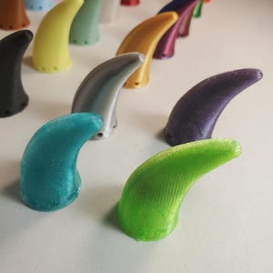 Op de afbeelding: Een verzameling kleurrijke, gebogen, hoornvormige objecten. De hoorns zijn in verschillende kleuren, waaronder groen, blauw, geel en paars. De objecten zijn in rijen gerangschikt, waarbij sommige scherp zijn en andere wazig op de achtergrond. De hoorns lijken van plastic te zijn.