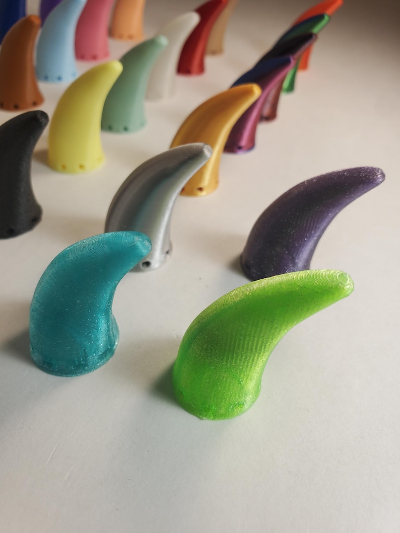 Op de afbeelding: Een verzameling kleurrijke, gebogen, hoornvormige objecten. De hoorns zijn in verschillende kleuren, waaronder groen, blauw, geel en paars. De objecten zijn in rijen gerangschikt, waarbij sommige scherp zijn en andere wazig op de achtergrond. De hoorns lijken van plastic te zijn.