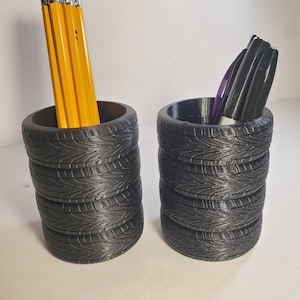 May include: Two black desk organizers shaped like stacked tires. One holds yellow pencils with pink erasers, the other holds black and purple markers. The organizers are on a white surface.