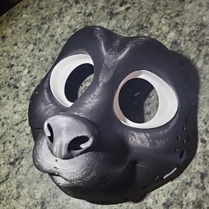 May include: A 3D-printed black animal mask featuring a prominent snout, a rounded nose, and two large, circular eye openings lined with white rims. The surface displays visible horizontal layer lines from the printing process and small ventilation holes.