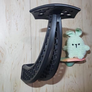 May include: A black, curved wall-mounted display stand featuring a structural honeycomb pattern throughout its frame. A small, light green crocheted plush creature with a simple embroidered face sits on a separate, small, multi-colored shelf nearby.