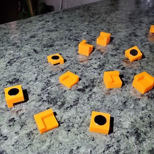 May include: An assortment of small, bright orange, 3D-printed geometric shapes. Some have a black circular detail. The shapes are scattered on a speckled gray surface. The objects appear to be components of a larger item.