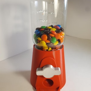 May include: A red gumball machine with a clear glass jar filled with colorful candies. The machine has a silver-colored dispensing mechanism and a small tray at the bottom. The jar has measurement markings.
