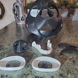 May include: A black, 3D-printed mask with separate components, including a nose, teeth, and eye frames. The mask has a rounded shape with several holes. Additional components are white and gray.