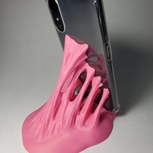 May include: A pink 3D printed phone stand with a textured, organic design. A black smartphone in a clear case is resting in the stand.