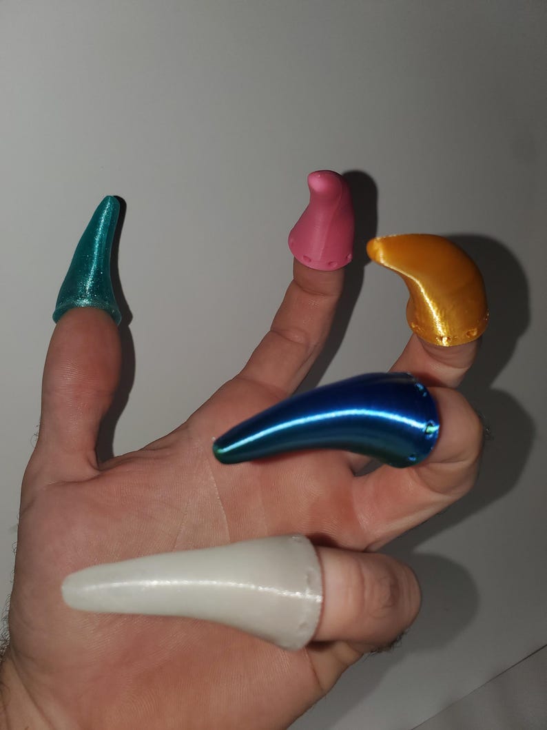 Op de afbeelding: Een hand met zes verschillend gekleurde vingernagels. De nagels zijn in tinten blauw, roze, goud en wit. De nagels zijn gemaakt van een glanzend materiaal en zijn ontworpen om over de vingertoppen te passen.