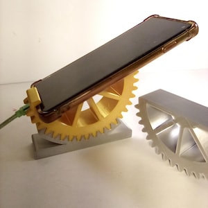 May include: A gold and silver phone stand with a gear design. The phone is black and is in a brown case. The stand is made of gold-colored plastic and silver metal. A green charging cable is plugged into the phone.