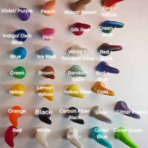 Op de afbeelding: Een display van kleine, gebogen objecten in verschillende kleuren, waaronder violet, perzik en houtbruin. De tekst "COLOR OPTIONS" staat bovenaan. Andere kleuren zijn rood, geel, blauw, groen en zwart. De objecten zijn gerangschikt in rijen en kolommen.