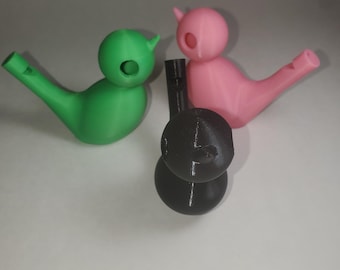 May include: Three 3D printed bird whistles in green, pink, and black. The whistles are shaped like birds with a small hole in the side for blowing.