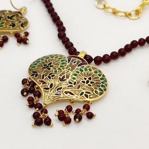 May include: A gold heart-shaped pendant necklace with a floral design and red beads. The necklace is made of gold and red beads. The pendant is decorated with green and black enamel.