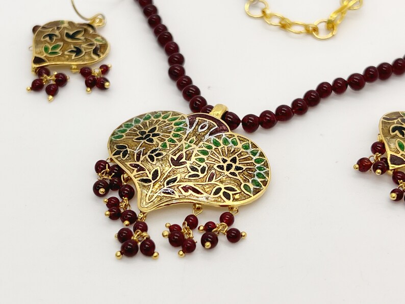 May include: A gold heart-shaped pendant necklace with a floral design and red beads. The necklace is made of gold and red beads. The pendant is decorated with green and black enamel.
