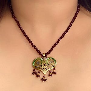 May include: A dark red beaded necklace with a gold pendant featuring a green and black floral design. The pendant is shaped like a heart and has small red beads dangling from the bottom.