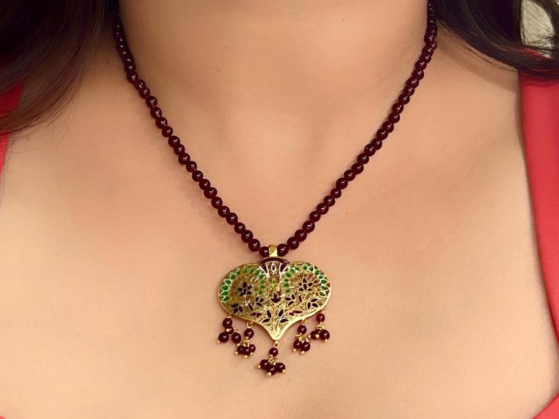 May include: A dark red beaded necklace with a gold pendant featuring a green and black floral design. The pendant is shaped like a heart and has small red beads dangling from the bottom.