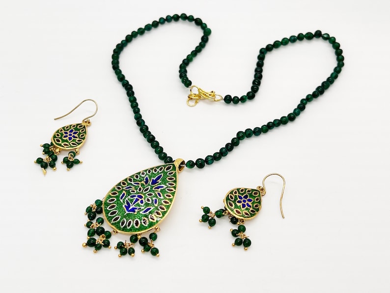 May include: A green gemstone necklace with a matching pair of earrings. The pendant and earrings feature a gold-toned teardrop design with intricate blue and green enamel work.