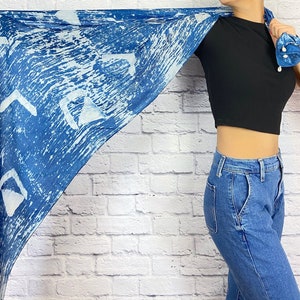 May include: A blue and white patterned scarf with a white background and blue abstract designs. The scarf is being held by a person wearing a black crop top and blue jeans.