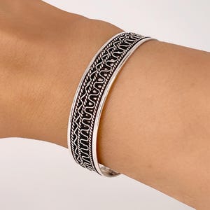 May include: A silver bangle bracelet with a detailed, intricate design. The bracelet is open and has a simple, elegant look.