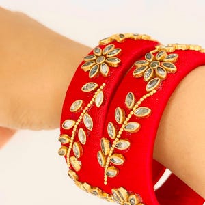 May include: Two red silk bangles with gold and silver embellishments. The bangles are decorated with floral and leaf designs.