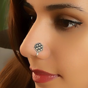 May include: A silver nose ring with a floral design. The ring is worn in the left nostril of a person with dark hair.