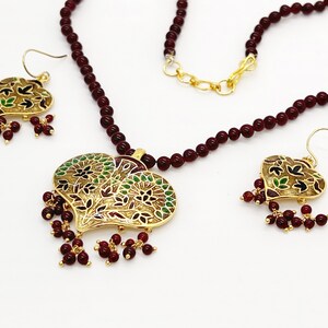 May include: A maroon beaded necklace with a gold heart-shaped pendant featuring intricate floral designs in green, black, and red enamel. The pendant is accented with maroon beads. Matching earrings with similar designs are also included.