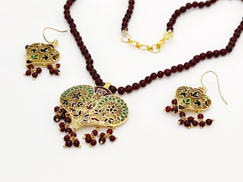 May include: A maroon beaded necklace with a gold heart-shaped pendant featuring intricate floral designs in green, black, and red enamel. The pendant is accented with maroon beads. Matching earrings with similar designs are also included.