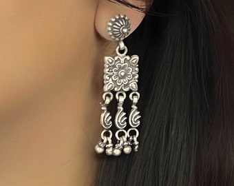May include: Silver dangle earrings with a square-shaped center piece featuring an intricate floral design. The earrings have three dangling chains with decorative elements and small silver beads at the bottom.