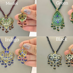 May include: Four sets of gold necklaces and earrings with enamel pendants. The pendants are in the shape of a heart and a teardrop. The necklaces are on a maroon, blue, green, and black background. The earrings are on a hand.