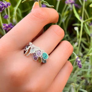 May include: A silver ring with multiple colorful gemstones, including turquoise, amethyst, and green stones. The ring is worn on a hand holding a sprig of lavender.