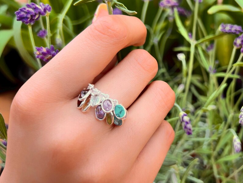 May include: A silver ring with multiple colorful gemstones, including turquoise, amethyst, and green stones. The ring is worn on a hand holding a sprig of lavender.