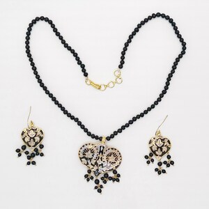 May include: A black beaded necklace with a gold heart pendant featuring intricate floral designs. The pendant has black and white enamel detailing. Matching earrings with the same design are included.