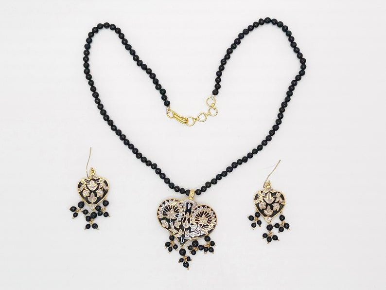 May include: A black beaded necklace with a gold heart pendant featuring intricate floral designs. The pendant has black and white enamel detailing. Matching earrings with the same design are included.