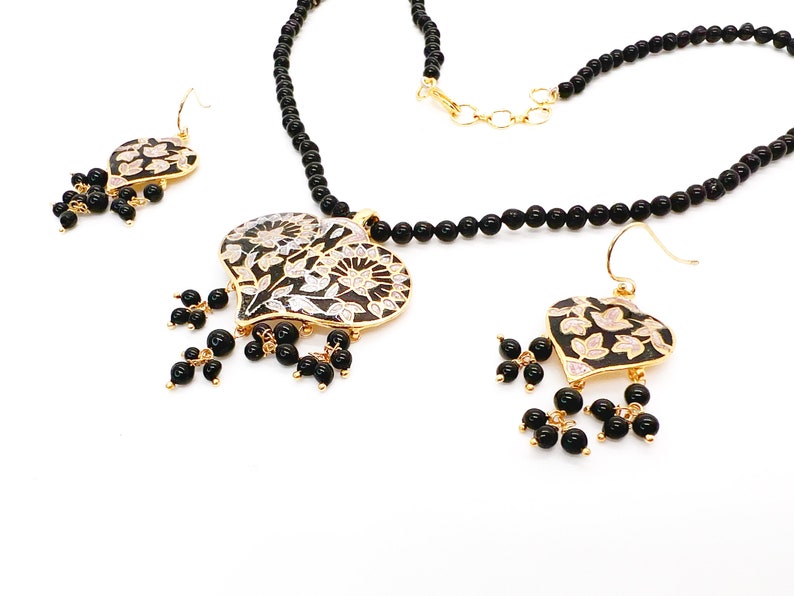May include: A black beaded necklace with a gold heart-shaped pendant featuring a floral design. The pendant is accented with black beads. Matching earrings with a similar design are included.