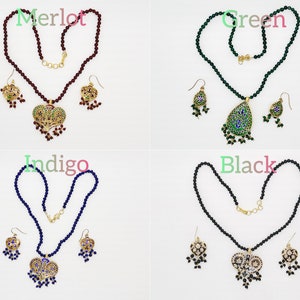 May include: Four sets of jewelry featuring a necklace and earrings. Each set has a different colored bead necklace and a pendant with a heart or teardrop shape. The pendants are decorated with intricate designs and colorful enamel. The colors of the necklaces are merlot, green, indigo, and black.