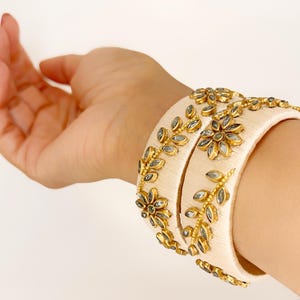Puede incluir: Dos brazaletes de seda marfil con adornos de diamantes de imitación dorados y transparentes en un diseño floral.