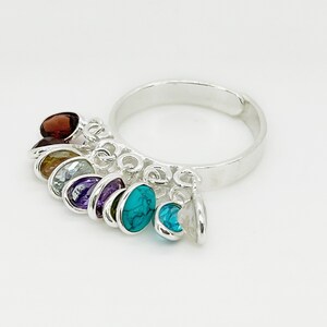 May include: A silver ring with a chain of dangling charms featuring different colored gemstones. The gemstones include red, orange, blue, purple, and turquoise.
