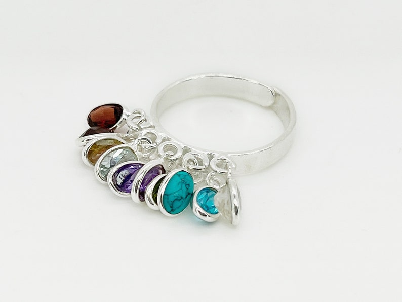 May include: A silver ring with a chain of dangling charms featuring different colored gemstones. The gemstones include red, orange, blue, purple, and turquoise.
