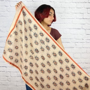 May include: A cream-colored scarf with a repeating geometric pattern in black and orange. The scarf has a red border.