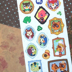 Puede incluir: Una hoja de pegatinas con varios personajes animales de la serie de videojuegos Animal Crossing. Las pegatinas son de varios colores y presentan diferentes poses y expresiones. Las pegatinas están sobre un fondo blanco y están rodeadas de un papel estampado en marrón y blanco.
