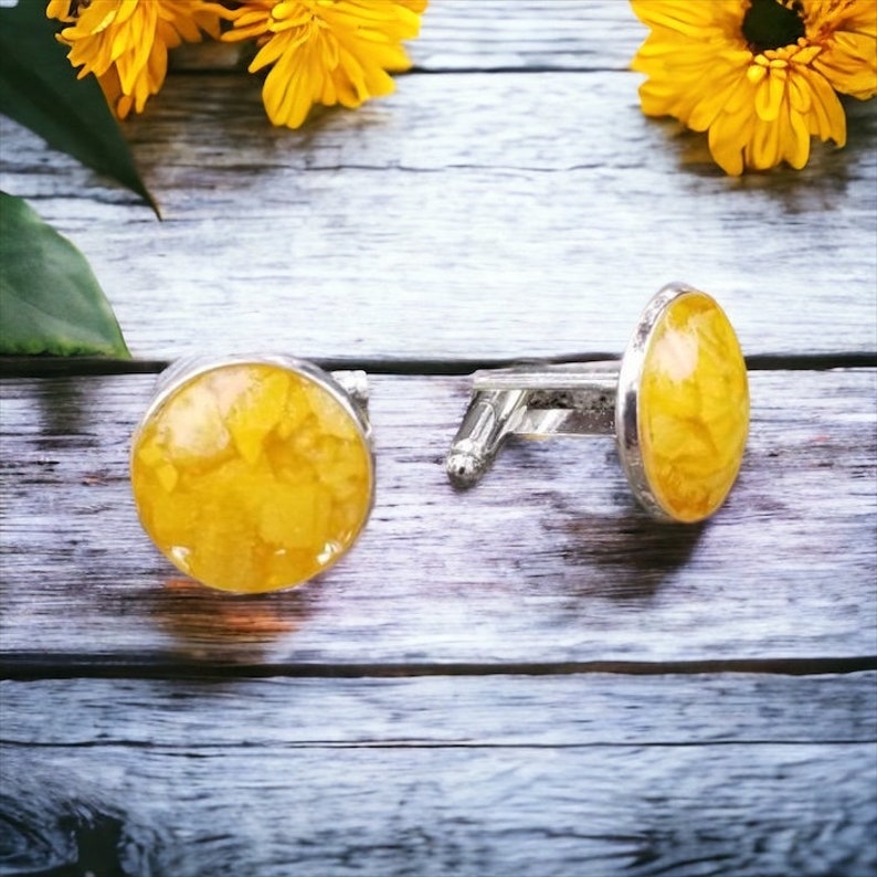 May include: A pair of silver cufflinks with yellow flower resin inserts. The cufflinks are on a light wood surface.