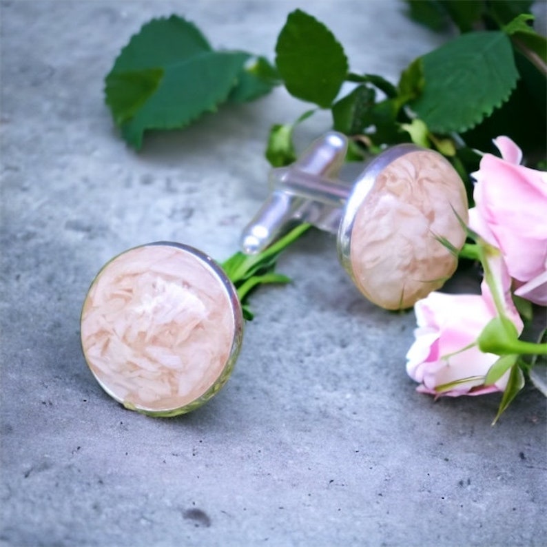 May include: A pair of silver cufflinks with pink and white marbled resin inserts. The cufflinks are surrounded by green leaves and pink flowers.