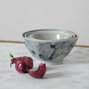 May include: A set of three nesting bowls with a white base and blue, green, and gray abstract patterns. The bowls are stacked on top of each other and are sitting on a white surface.
