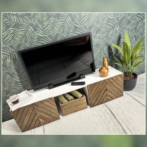 May include: A white TV stand with a wood grain finish and two drawers. A flat screen TV is on top of the stand, and there are two gold vases and a potted plant on the shelf.