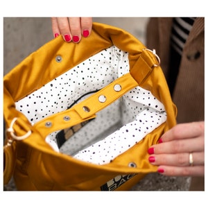 May include: Mustard yellow tote bag with a white interior featuring black polka dots. The bag has a snap closure and a strap. The bag is open, revealing the interior. The bag is held open by a person.