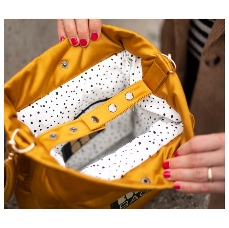 May include: Mustard yellow tote bag with a white interior featuring black polka dots. The bag has a snap closure and a strap. The bag is open, revealing the interior. The bag is held open by a person.