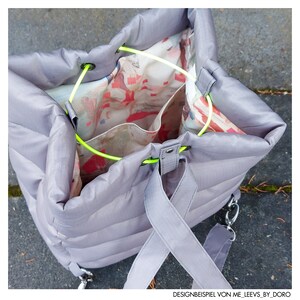 May include: A grey, quilted tote bag with a drawstring closure and neon green accents. The interior features a colourful floral pattern. The bag has a shoulder strap and a silver chain detail. The text "DESIGNBEISPIEL VON ME_LEEVS_BY_DORO" is visible.