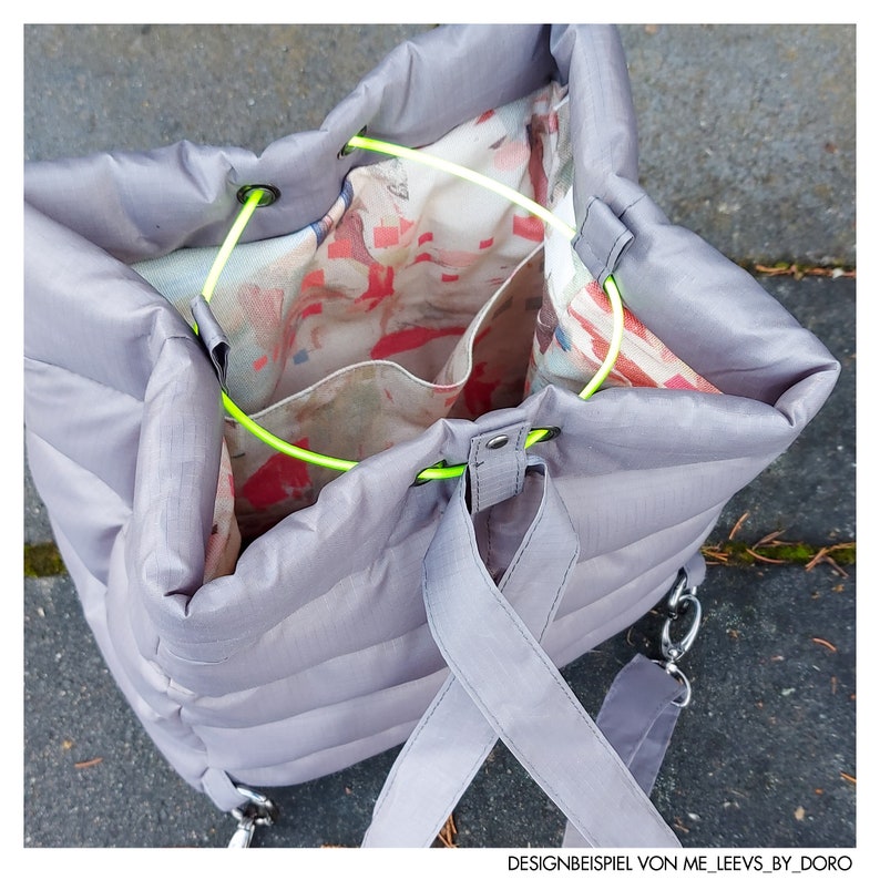 May include: A grey, quilted tote bag with a drawstring closure and neon green accents. The interior features a colourful floral pattern. The bag has a shoulder strap and a silver chain detail. The text "DESIGNBEISPIEL VON ME_LEEVS_BY_DORO" is visible.