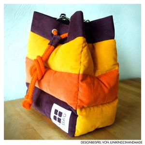 May include: A corduroy bag in a vibrant colour scheme of purple, orange, and yellow horizontal stripes. It features a drawstring closure with an orange rope and a small white tag with a logo. The bag is placed on a wooden surface.