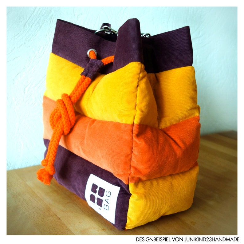 May include: A corduroy bag in a vibrant colour scheme of purple, orange, and yellow horizontal stripes. It features a drawstring closure with an orange rope and a small white tag with a logo. The bag is placed on a wooden surface.