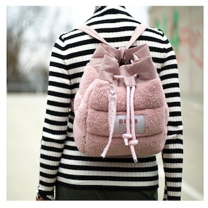 May include: A pink, quilted backpack with a drawstring closure and a fluffy, textured exterior. The backpack has a small, rectangular tag with white text. The straps and drawstring are a lighter shade of pink.